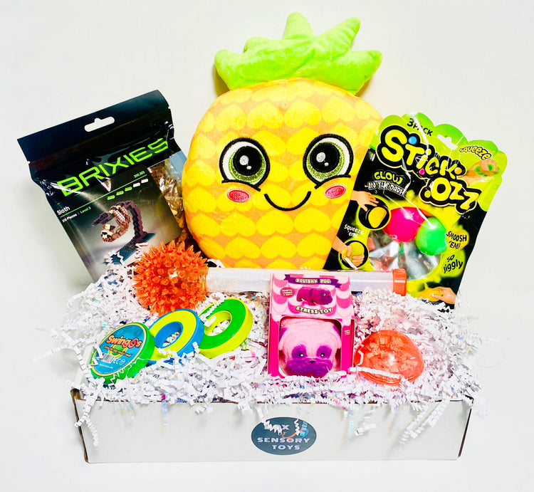 A child's gift box filled with colorful toys and treats, featuring a large plush, sticky squish balls,  Brixies, glow wand, swings magnets slide 3-ring fidget, stress toy, and other sensory toys in white box with shredded paper.